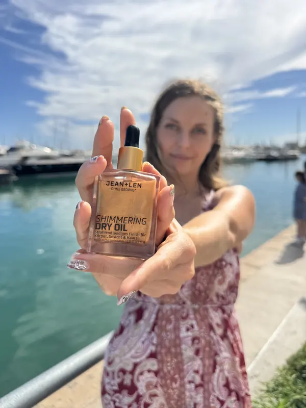 Jasmin präsentiert ein schimmerndes Körperöl vor einem Yachthafen